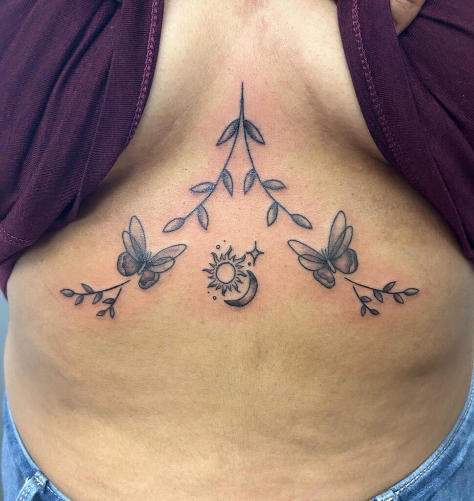 Under-breast tattoo with butterflies, leafy branches, and celestial symbols including a sun and crescent moon