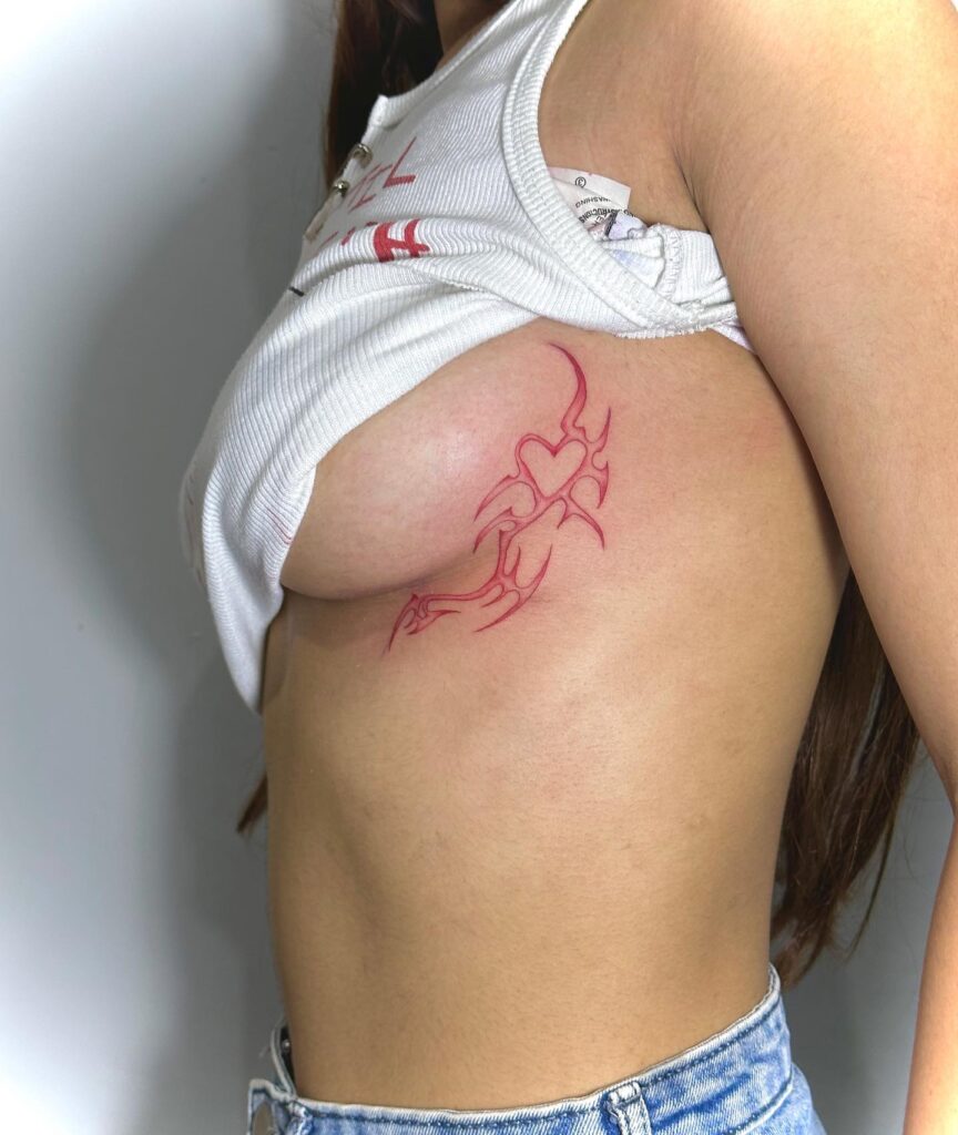 Side under-breast tattoo in red ink featuring a tribal heart design with sharp, flowing lines