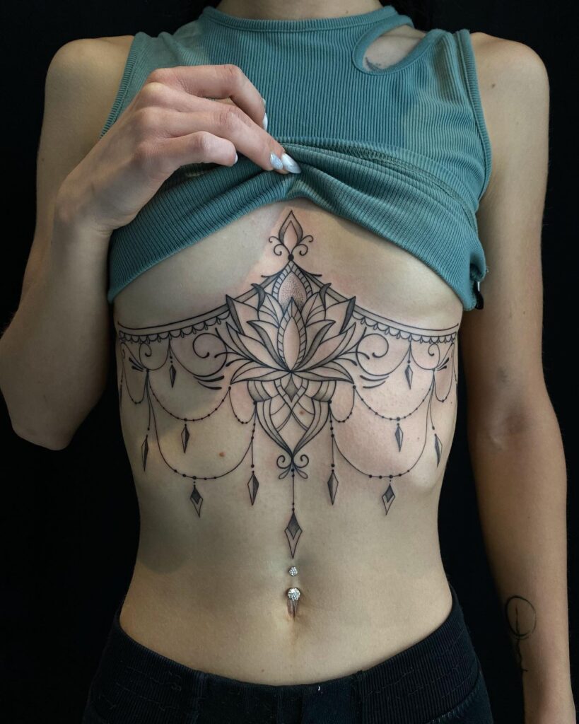 Under-breast tattoo featuring an intricate lotus with mandala patterns and elegant chain accents, creating a chandelier effect.