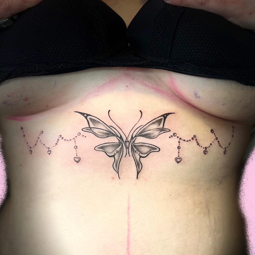 Under-breast tattoo featuring a butterfly with shaded wings and heart chain details, creating a feminine and delicate design