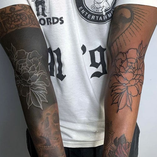 Two forearm tattoos featuring a blackout sleeve with white ink peony designs on one arm and a traditional black ink peony tattoo on the other