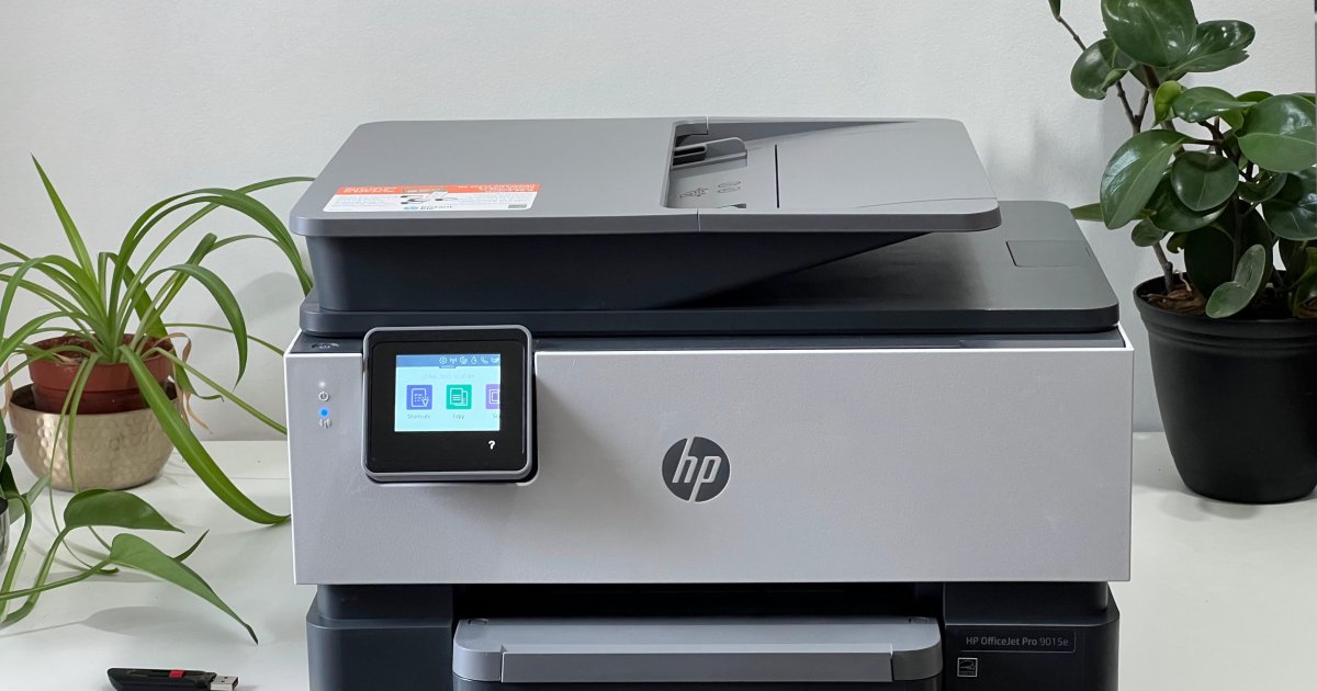 An HP OfficeJet Pro 9015e all in one printer rests on a white table with plants and a thumb drive beside it