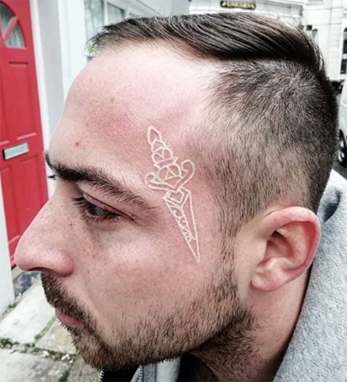 A white ink tattoo featuring a detailed dagger with a rose entwined around it, placed on the side of the face