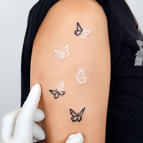 A collection of minimalist white and black butterfly tattoos scattered across the upper arm, featuring a mix of outline designs