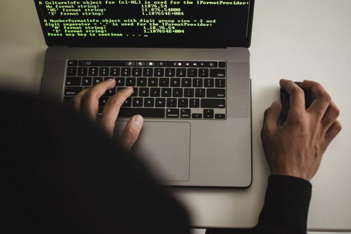 A hacker is typing on an Apple MacBook laptop, showing code on his screen.