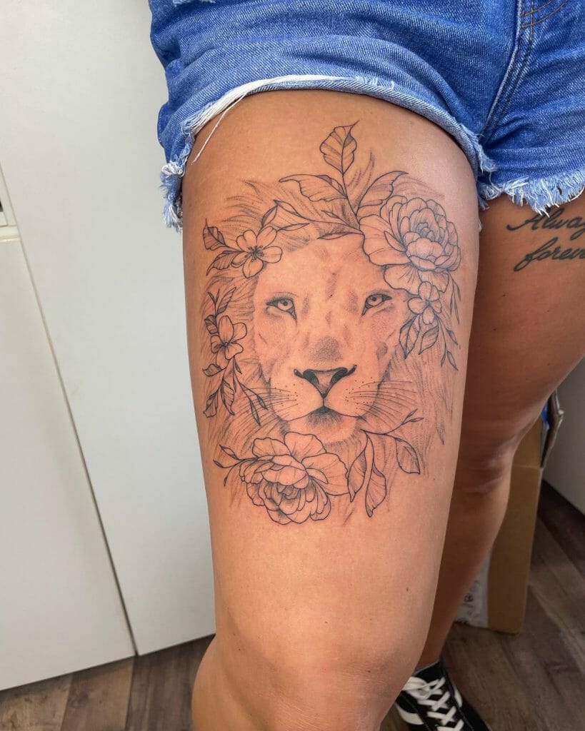 Thigh tattoo of a lion's face surrounded by delicate flowers and leaves, designed with soft shading and fine lines.