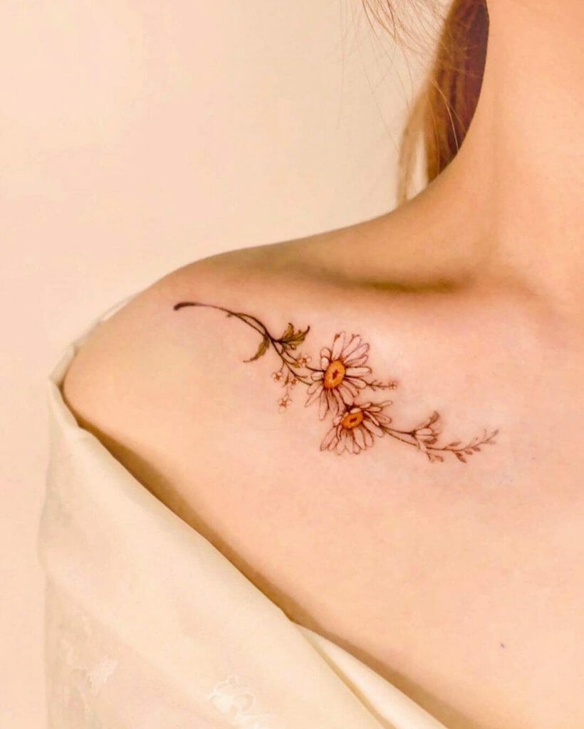 A delicate tattoo of two daisies with thin stems and leaves in soft orange and brown tones, placed along the collarbone