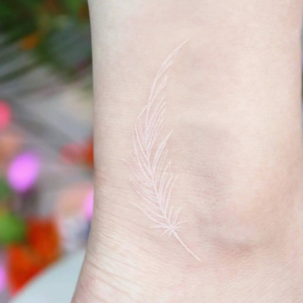 A minimalist white ink feather tattoo placed on the ankle, showcasing fine line work and soft detailing