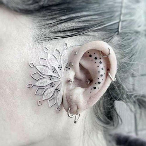 A white ink mandala tattoo extending from behind the ear with black dot accents along the ear, creating an intricate design