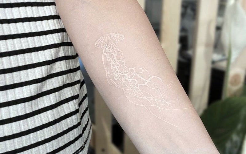 A minimalist white ink jellyfish tattoo featuring delicate, flowing tentacles and a soft design, placed on the forearm