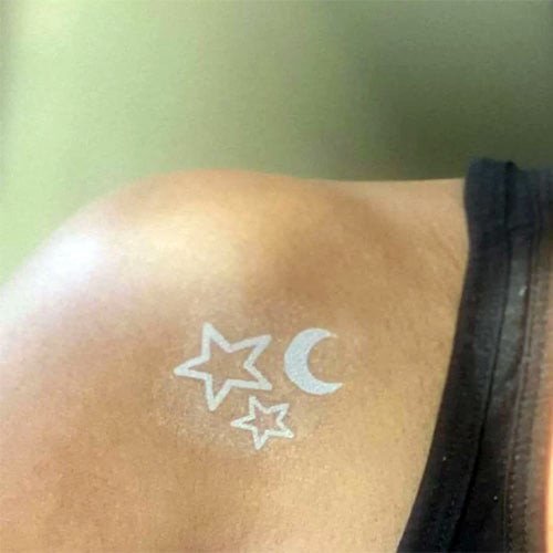 A minimalist white ink tattoo featuring two stars and a crescent moon placed on the shoulder