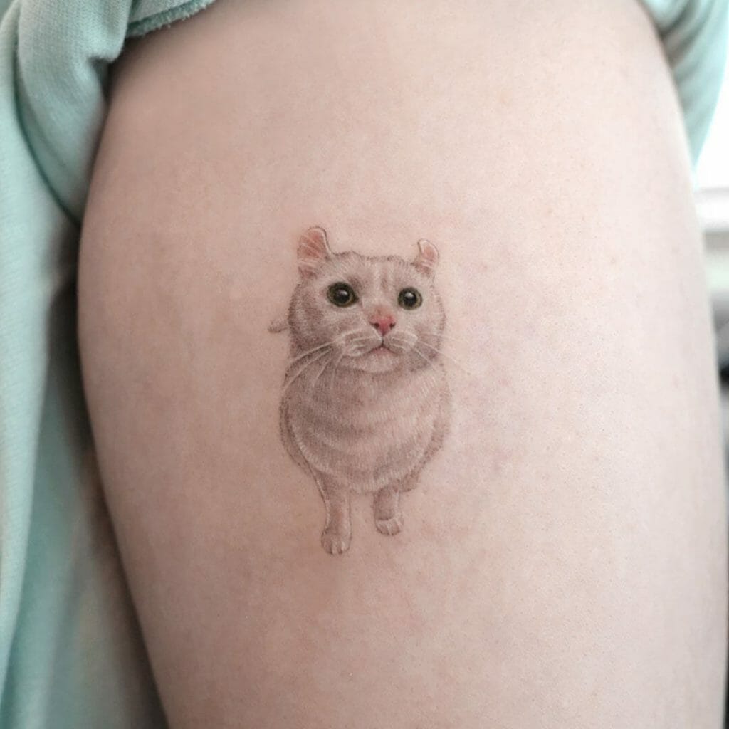 A small, realistic cat portrait tattoo featuring a light-colored cat with green eyes and pink nose, inked on the upper arm