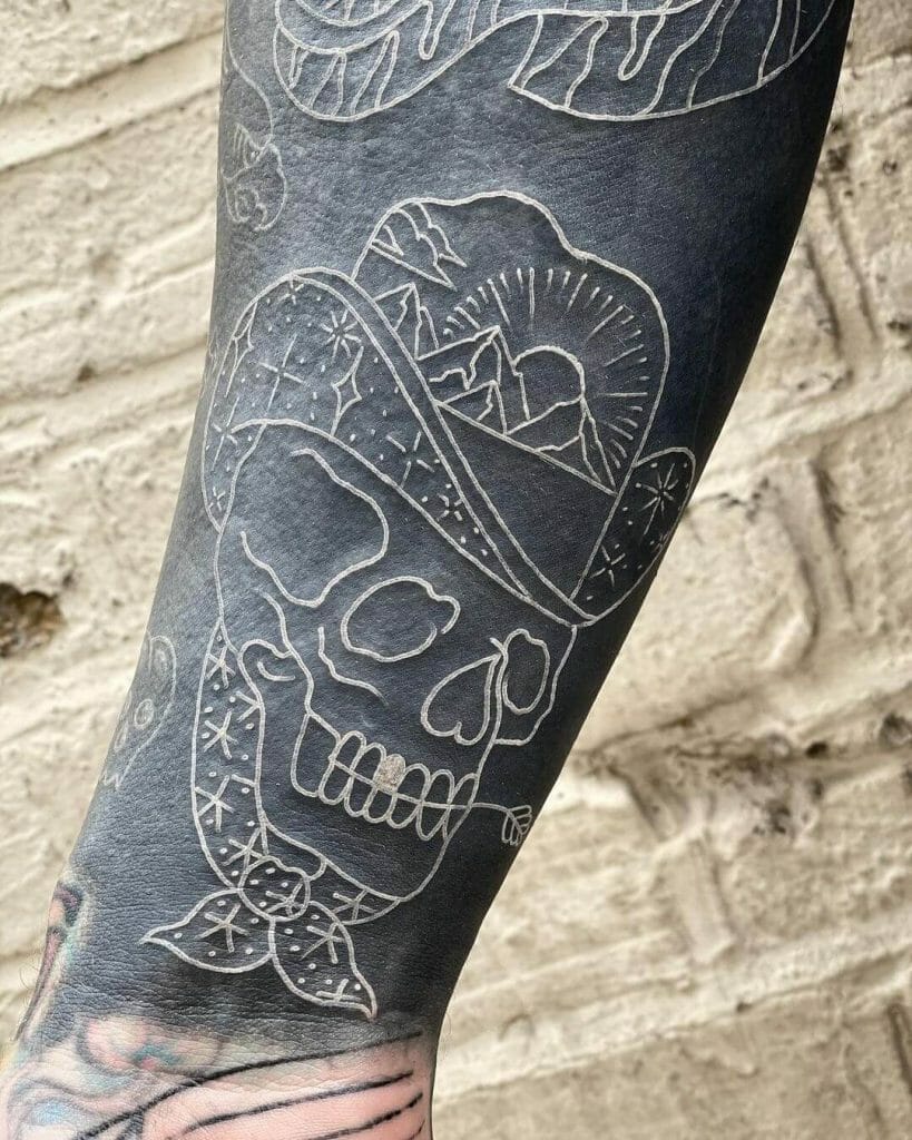 A white ink tattoo featuring a cowboy skull wearing a hat with a mountain scene, placed on a blacked-out sleeve background