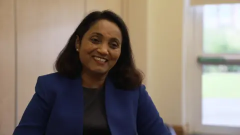 Geeta Kumar is a woman with shoulder-length dark hair. She is wearing a blue jacket and black top and is smiling. She is sitting in an office with a window in the background.