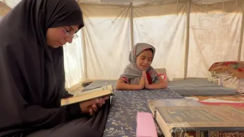 A woman reads the Quran with her daughter at a table inside a tent 