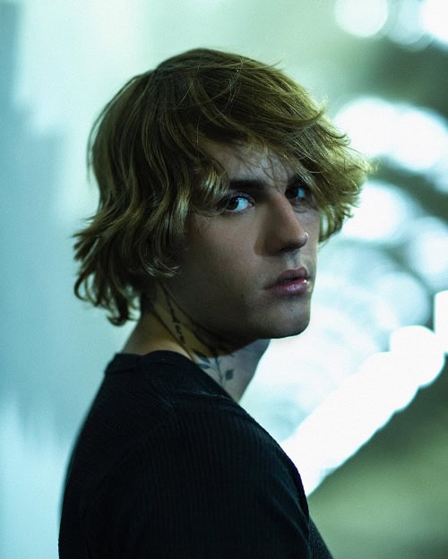 A man with tousled medium-length hair, wearing a black shirt, standing in moody, soft lighting with a contemplative expression.