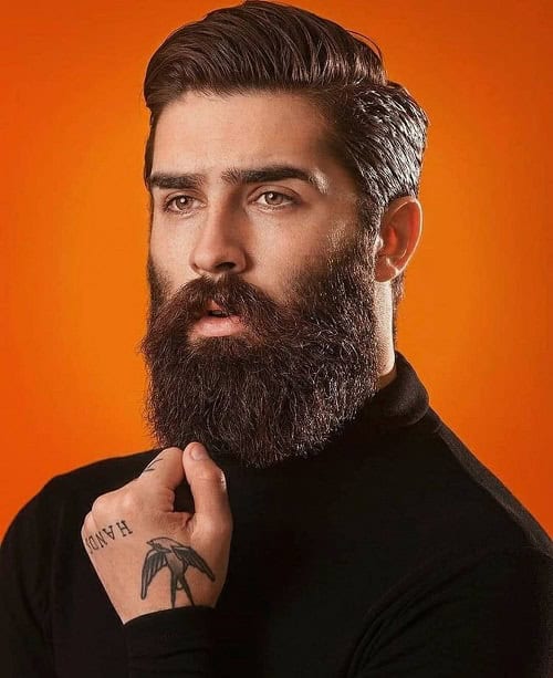 Man with a slicked-back crew cut hairstyle and full beard, wearing a black sweater against an orange background