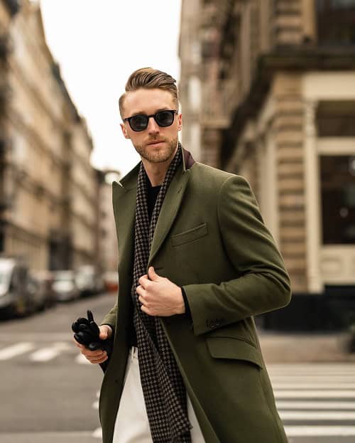 A man with a slicked-back hairstyle, wearing sunglasses, a green overcoat, and a scarf, walking confidently down a city street while holding an umbrella.