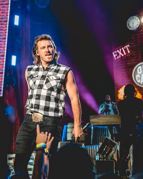 Man on stage with a country mullet haircut, featuring a side part and long back, styled in a rugged look