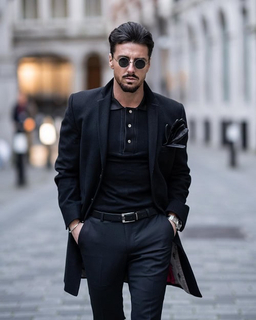 A man with slicked-back hair, wearing a black coat and sunglasses, walking down a city street in a sleek, all-black outfit, showcasing a modern and sophisticated street style look.