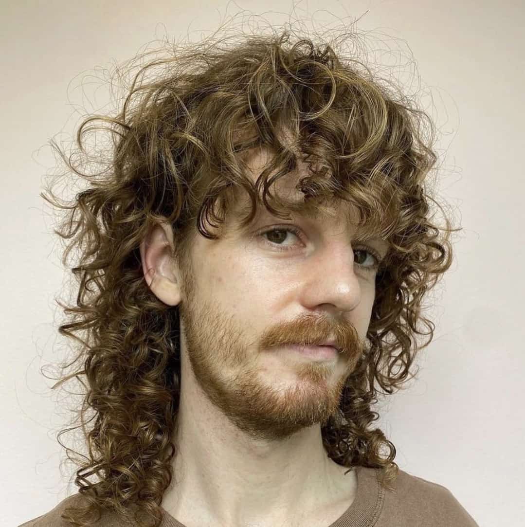 Front and side view of a man with a long curly mullet haircut, featuring thick curls cascading down the back and textured bangs in the front