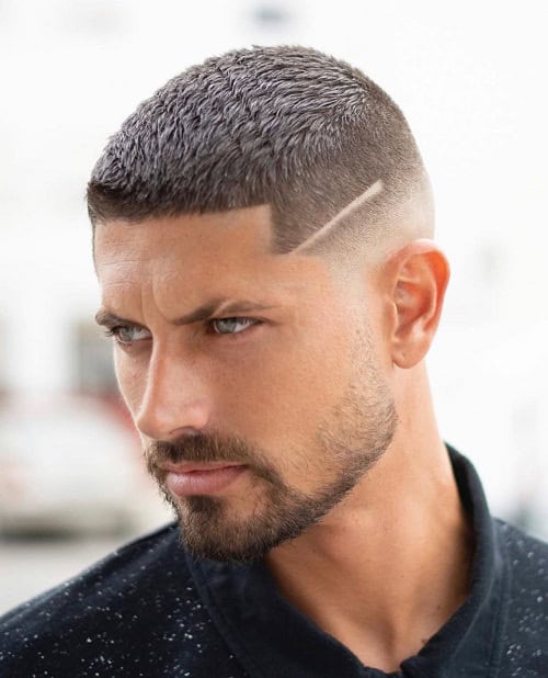 Man with a sharp crew cut hairstyle featuring a line design on the side and a faded look, paired with a beard.