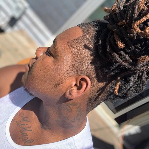 Man with dreadlocks tied up and a crew cut fade on the sides, wearing a white tank top outdoors.