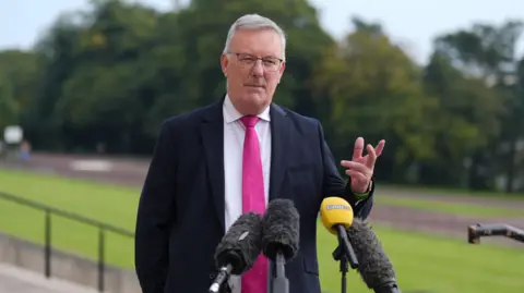 PA Mike Nesbitt standing behind four microphones while speaking to the media. He is wearing a dark suit with a white shirt and a pink tie. 