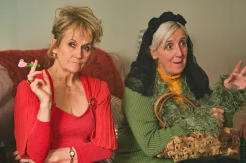 BBC/Expectation TV/Matt Squire Lorraine Ashbourne (Joan) with Aunty Ange (Julie Hesmondhalgh) - Lorraine is wearing a red low-cut top and Ange has a black veil on her head and is wearing a yellow top and green cardigan