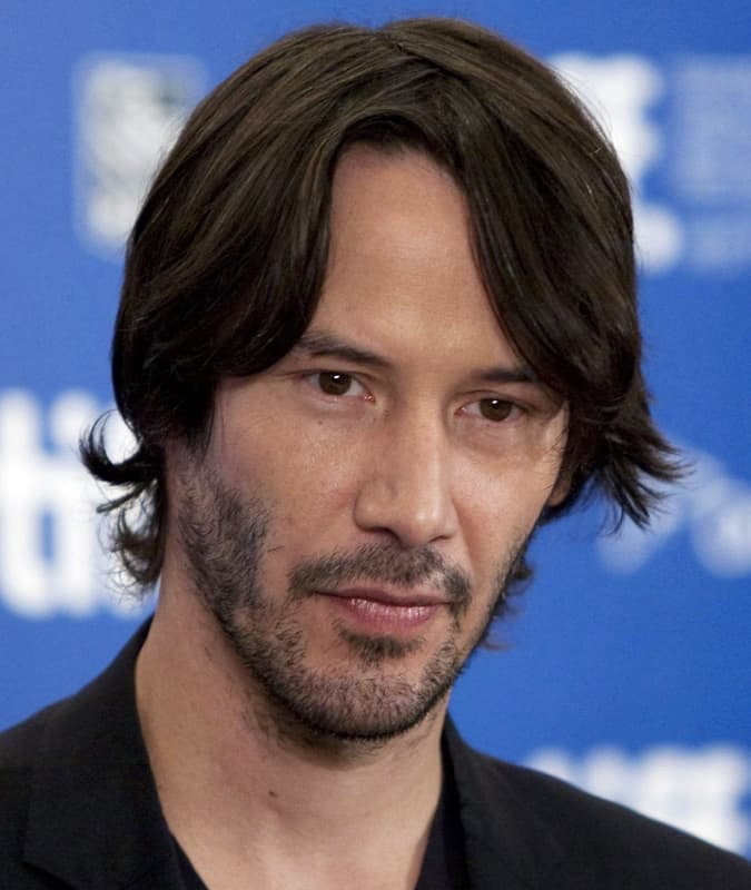 Keanu Reeves with a modern Eboy haircut featuring natural layers and a subtle middle part, wearing a dark blazer.