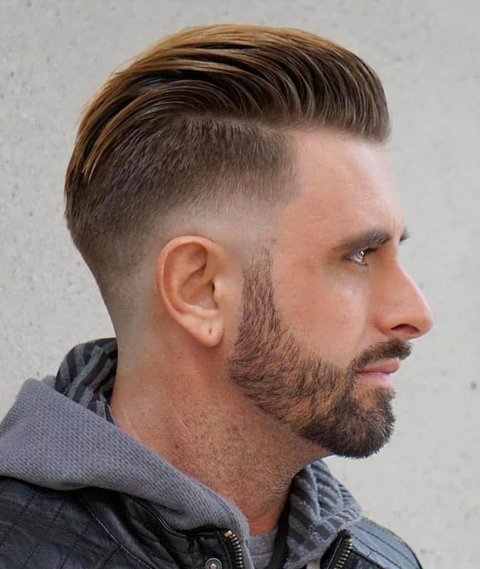 Side profile of a man with a low drop fade haircut, slicked back top, and neatly trimmed short beard.