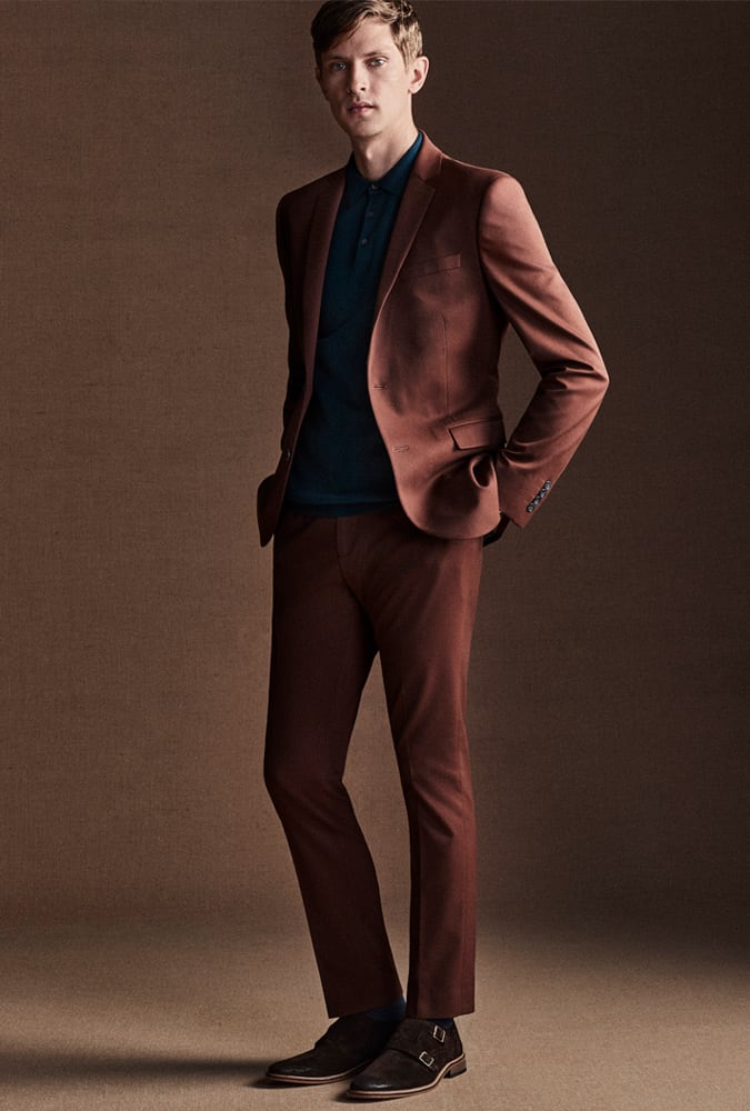 Man in a tailored rust-colored suit with a dark teal polo shirt and brown double-monk strap shoes, exuding a refined and modern style