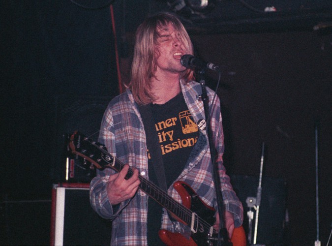 Kurt Cobain, 1990 - Kurt Cobain performing on stage wearing a plaid flannel shirt and graphic tee, holding a guitar and singing into a microphone