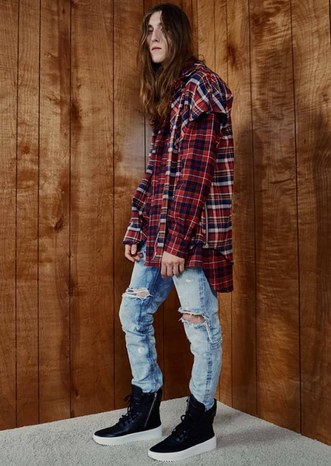 Young man in a layered grunge outfit with an oversized plaid shirt, distressed jeans, and black high-top sneakers, standing against a wooden wall