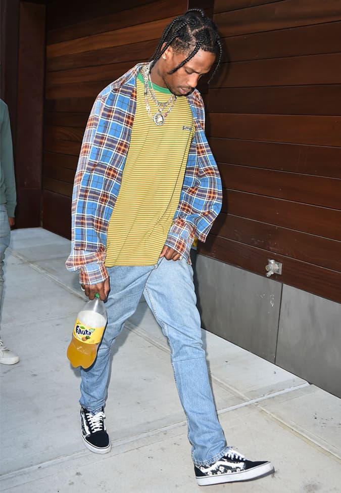 Travis Scott wearing a layered grunge street style outfit with a plaid shirt over a striped tee, light-wash jeans, and black sneakers