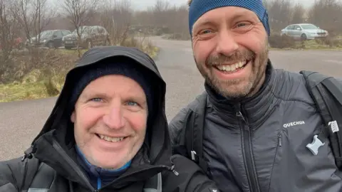 A selfie with Glenn and his friend Nikolaj in front of a car park before they started the climb. Both men are dressed in black jackets and are smiling at the camera.