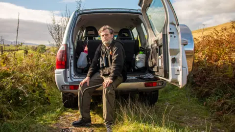 Dafydd Hughes holds a whip and looks into the camera. He is sat on the ledge of his vehicle with the boot door open. 