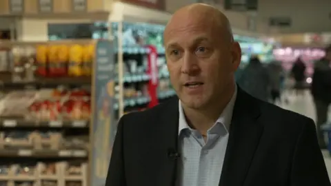 Lidl UK boss Ryan McDonnell wearing a black blazer and white shirt with a pale blue small check standing in the supermarkets with aisles and shoppers in the background