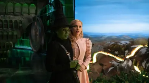 Universal Elphaba dressed in a black hat and dress has a smirk on her face whilst Glinda, dressed all in pink, appears surprised.