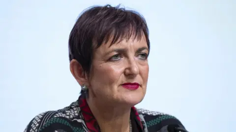 PA Media Angela Constance, with dark brown hair, sits in front a light blue background 