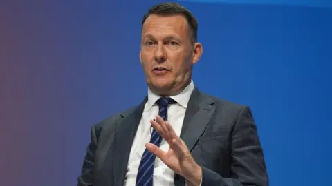 PA Media Russell Findlay, wearing a dark blue suit and tie with a white shirt, speaks with his left hand in front of body in front a blue background 