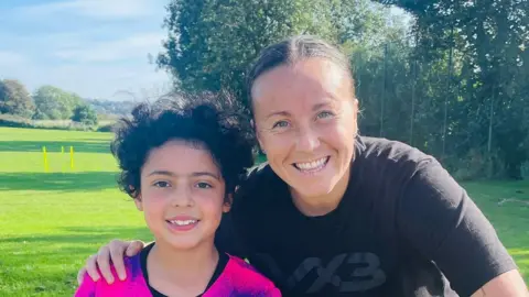 Wilmar Manzano Natasha Allen-Wyatt poses with her arm around Elen on a grass football pitch. Elen has dark brown, curly hair and is wearing a pink and blue football shirt. Ms Allen-Wyatt has her hair tied back and is wearing a black training top.