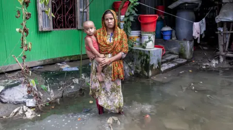 Muhammad Amdad Hossain/Getty Images A woman holds a small child in her arms in front of her flooded house in a village in Bangladesh