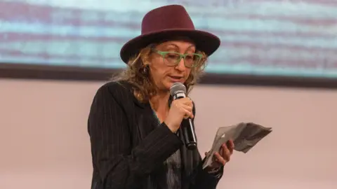 EPA Halyna Hutchins' friend Rachel Manson reads from a piece of paper. She's wearing a red hat and speaks into the microphone.