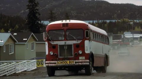 Getty Images A red school bus driving on a residential road with a lake and forest in the background, photographed in Saskatchewan's Uranium City in 1975. 