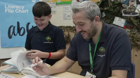 Gemma Laister/BBC Jacob, wearing a navy polo shirt with the Perryfields logo on it, smiles as he turns the page of a workbook. He is sitting next to a member of staff who has grey hair and is holding a black marker pen.