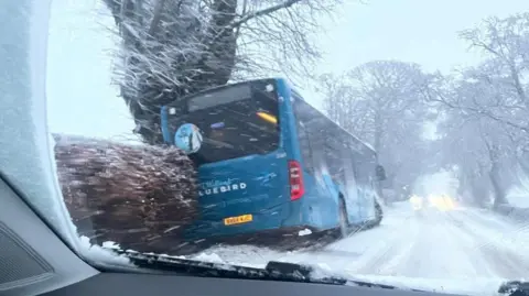 Facebook/Evelyn Tweed A blue bus is tilted to the side after going off the road. The photo is taken through a car windscreen.