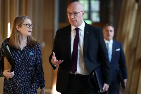 Getty Images Shirley-Anne Somerville (left) is walking, carrying a black document folder, beside John Swinney in a corridor at Holyrood. Alex Cole-Hamilton can be seen in the background over Swinney's shoulder.