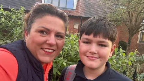 Jo Jo, who has brown hair tied back into a ponytail and sunglasses on her head, takes a selfie alongside her son Jacob. He has dark brown hair too. They're stood in front of a leafy green hedge.