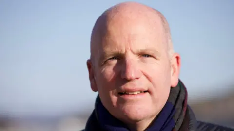 A close-up of Glenn Campbell looking directly at the camera, with a blue sky behind him. He is wearing a scarf and a black jacket.